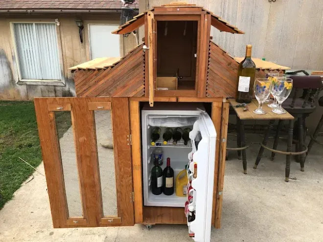A custom wooden bar cabinet with a roof-like structure, an open door, a small refrigerator inside, and wine glasses nearby.