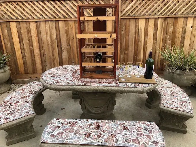 A mosaic stone table and benches in an outdoor setting, featuring a wooden wine rack, a wine bottle, and several glasses.