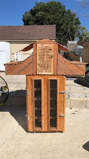 A homemade wooden structure with a decorative, slatted top section and a lower cabinet with two mesh-screened doors.