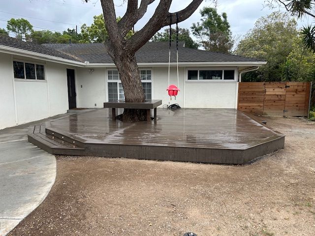 A dark brown wooden deck surrounds a large tree with a built-in bench and hanging swing in a residential backyard.