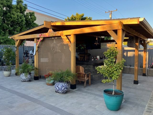A wooden outdoor patio cover with an attached bar area, surrounded by various potted plants on a stone paved surface.