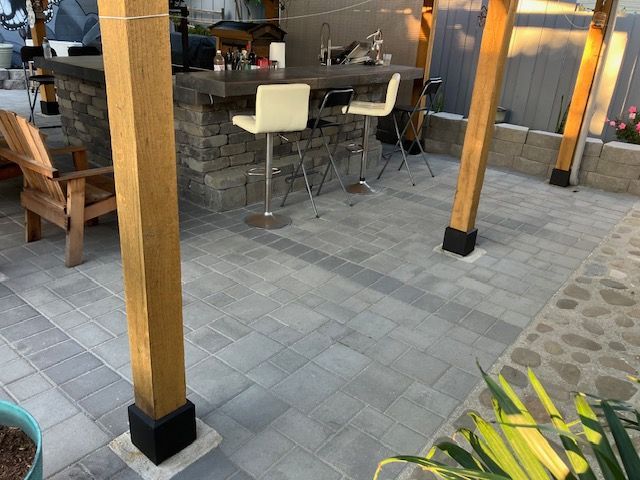 A backyard patio with a stone bar, wooden support beams, stools, and a wooden chair on stone pavers.