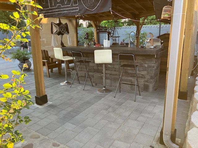 An outdoor bar with a stone counter and four stools under a wooden patio cover, set on a gray paver patio.