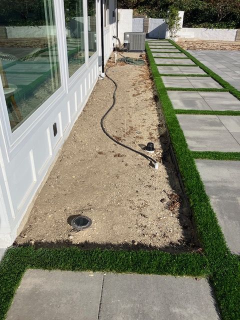 A narrow, gravel-filled dirt strip along a white building wall, adjacent to a paver and turf walkway.
