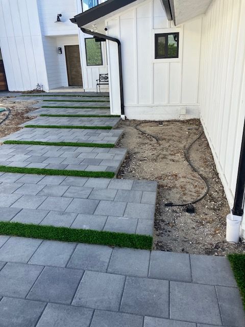 A walkway of large gray pavers with artificial turf gaps leads to a modern white house with a dirt patch beside it.