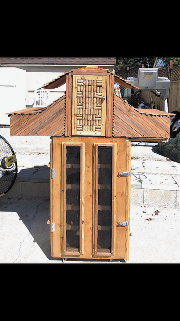 A vertical, wooden smoker or coop structure with a slanted roof, central door, and two narrow glass-paned doors below.