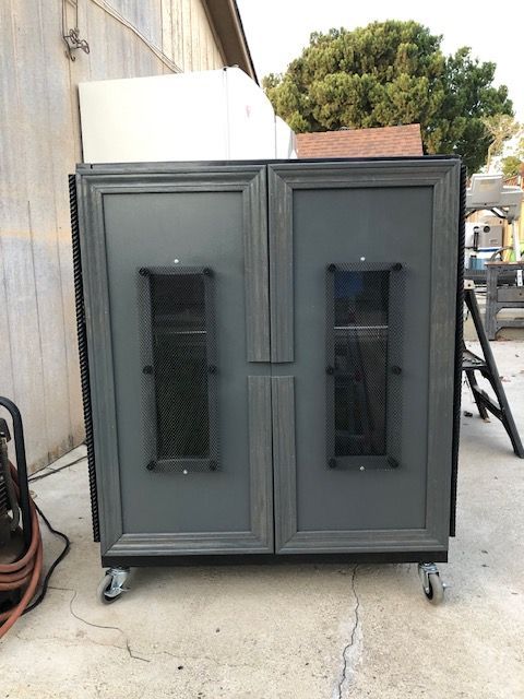 A grey, double-door metal equipment cabinet with rectangular viewing windows, set on wheels outdoors.