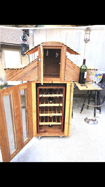 A wooden wine cabinet with a roof-like top and glass doors, standing outdoors with a wine bottle on an attached side table.