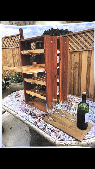 A wooden wine rack stands on a table outdoors next to a serving tray holding empty wine glasses and a bottle of wine.