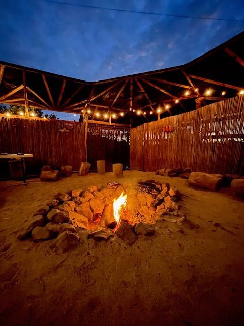 A fire pit with a small flame surrounded by rocks in a sandy, thatched-walled enclosure lit by string lights at dusk.