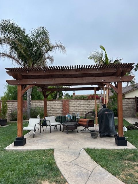 A wooden pergola sits on a concrete patio in a grassy backyard with patio chairs, a fire pit, and a grill cover.