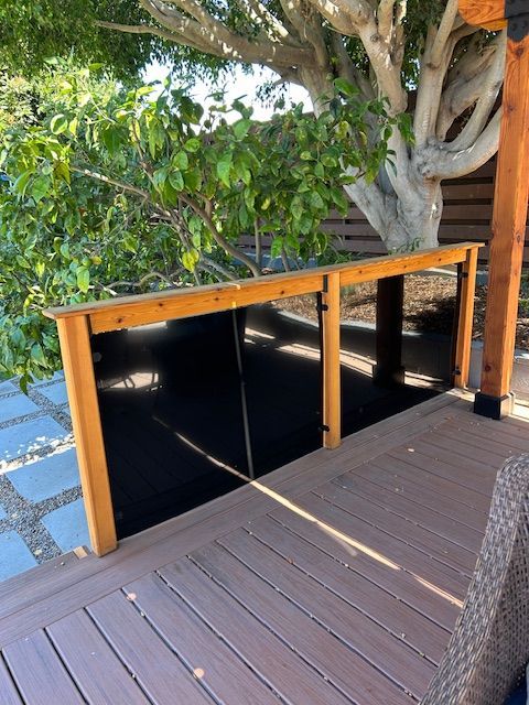 A wooden deck railing with dark glass panels stands beside a leafy tree and a patio area with pavers.