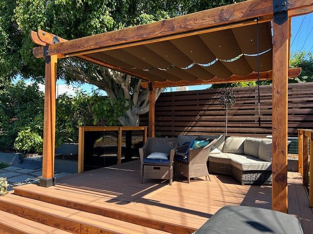 A brown wooden pergola with a retractable fabric canopy stands over an outdoor deck with patio furniture and a privacy fence.