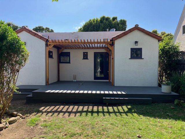 A backyard view of a white cottage with a wooden pergola, a dark gray deck, and a glass door on a sunny day.