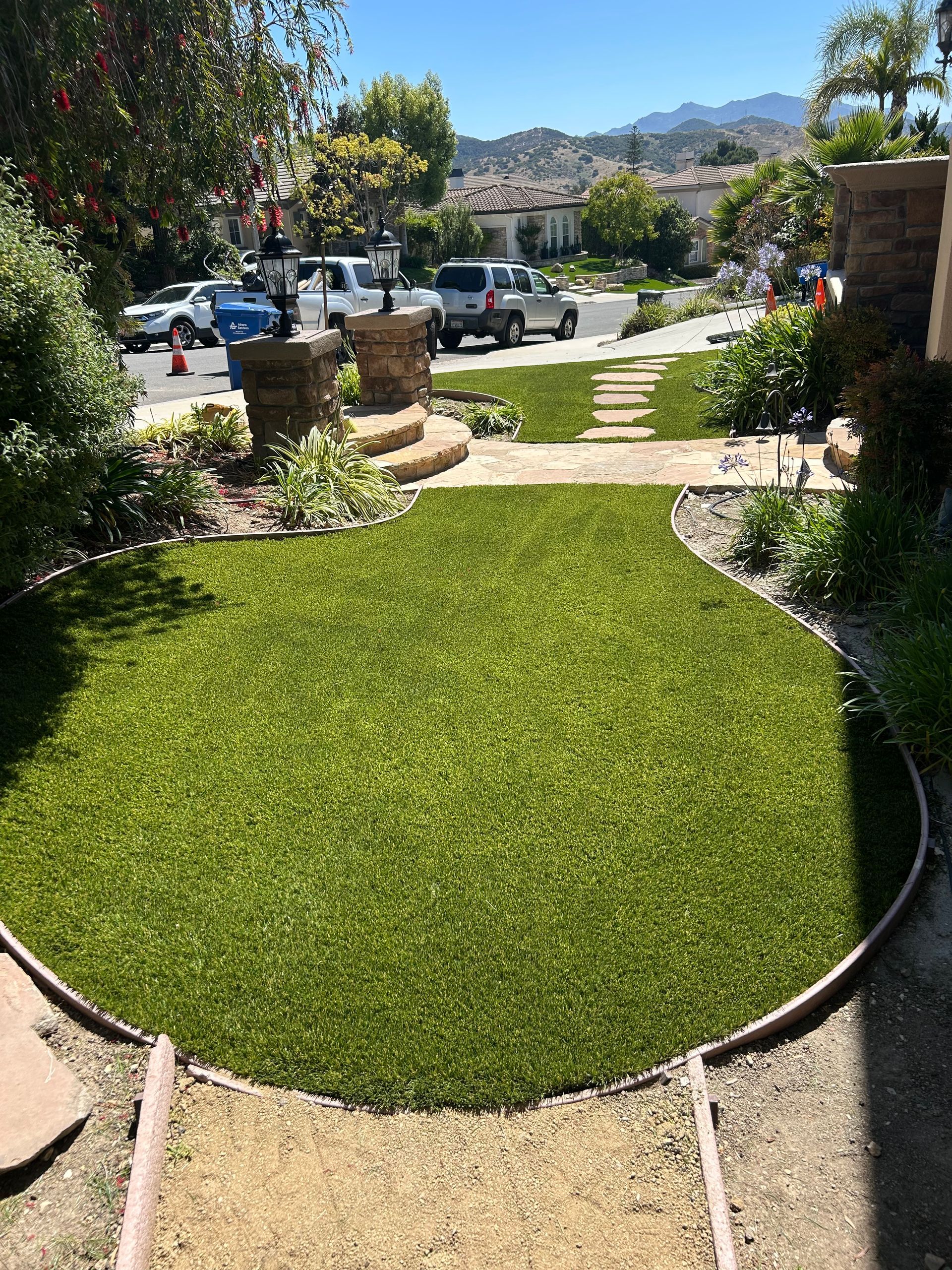 A gravel path with wooden steps winds through a landscaped garden featuring evergreen shrubs, small trees, and large rocks.