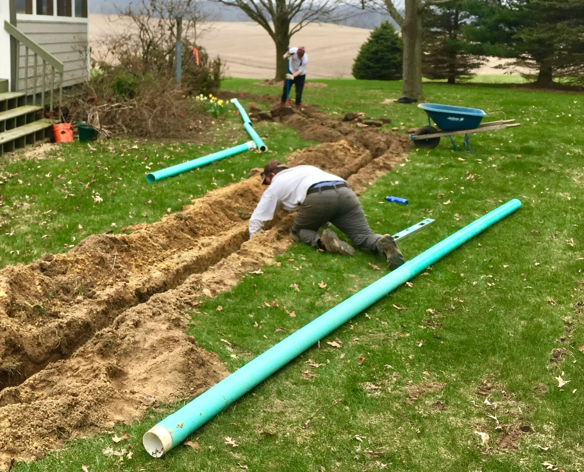 Two people in a grassy yard install green drainage pipes in a trench, with a wheelbarrow nearby.