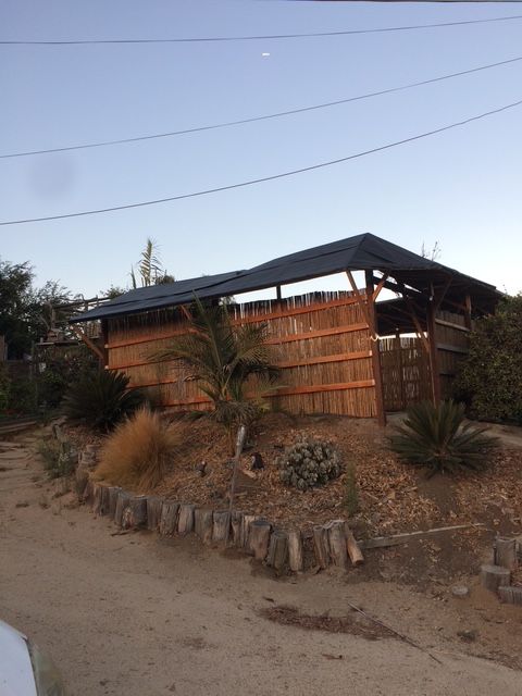 A rectangular shelter with a dark roof and walls made of vertical wooden slats, set on a sloped garden bed with greenery.