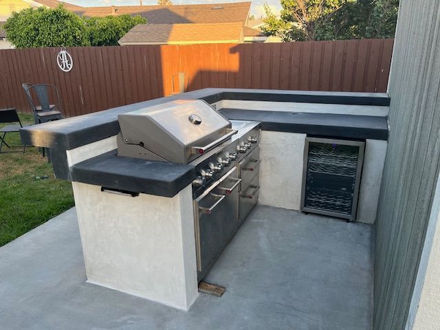 An outdoor kitchen island with a stainless steel grill, a small beverage fridge, and dark gray concrete counters.