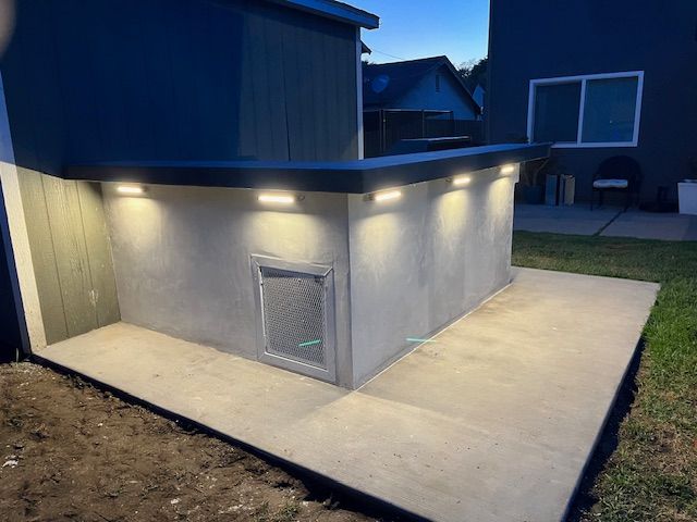 A concrete outdoor bar with built-in, illuminated wall-mount lights and a metal vent panel, situated on a patio at dusk.