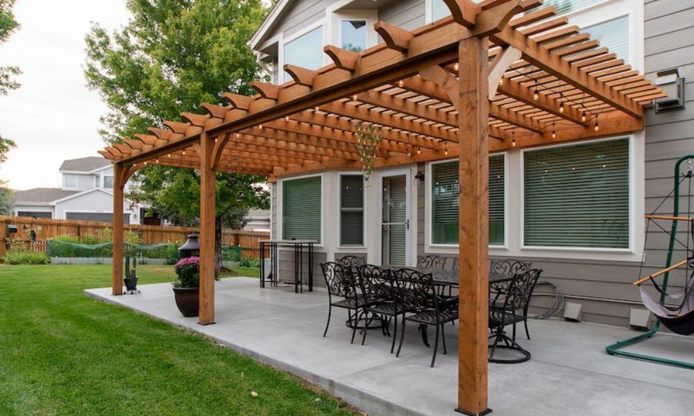 A wooden pergola covers a concrete patio with a black metal dining set and a hanging chair in a backyard.