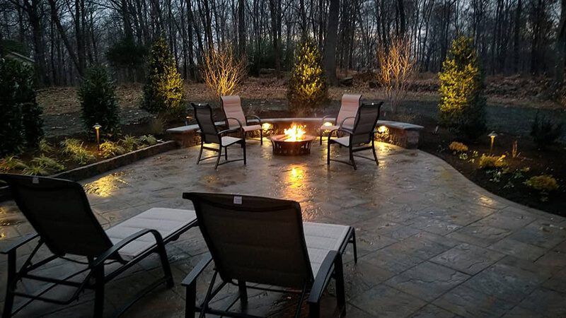 A stone patio at dusk with a lit fire pit, surrounded by patio chairs, landscaping, and warm accent lighting.