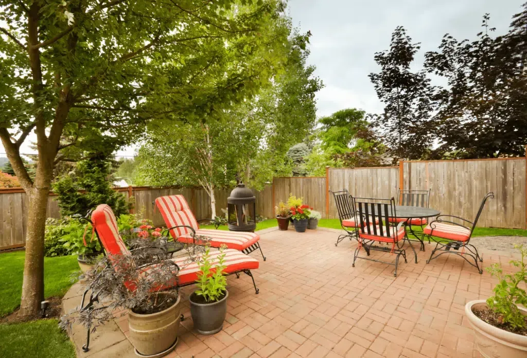 A brick patio in a backyard features lounge chairs with red striped cushions, a dining set, and potted plants near a fence.