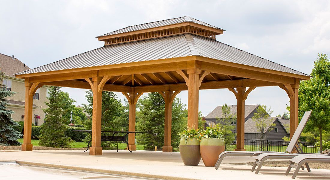 A wooden pavilion with a tiered roof sits on a patio with two planters and lounge chairs nearby.