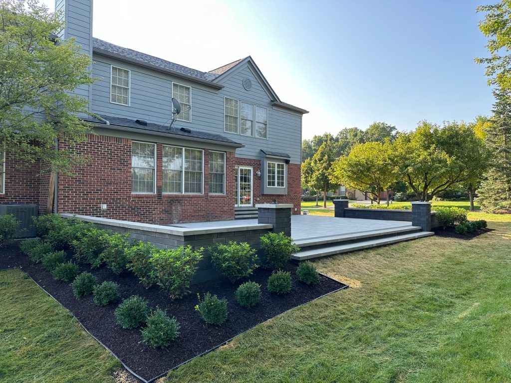 A brick house with grey siding features a newly constructed patio with stone steps and pillars surrounded by landscaping.