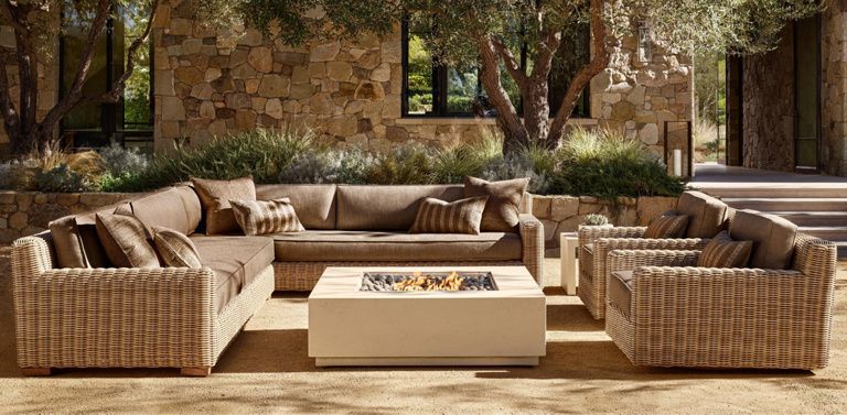 A tan wicker sectional sofa and matching chairs set around a square, white fire pit table on a paved outdoor patio.