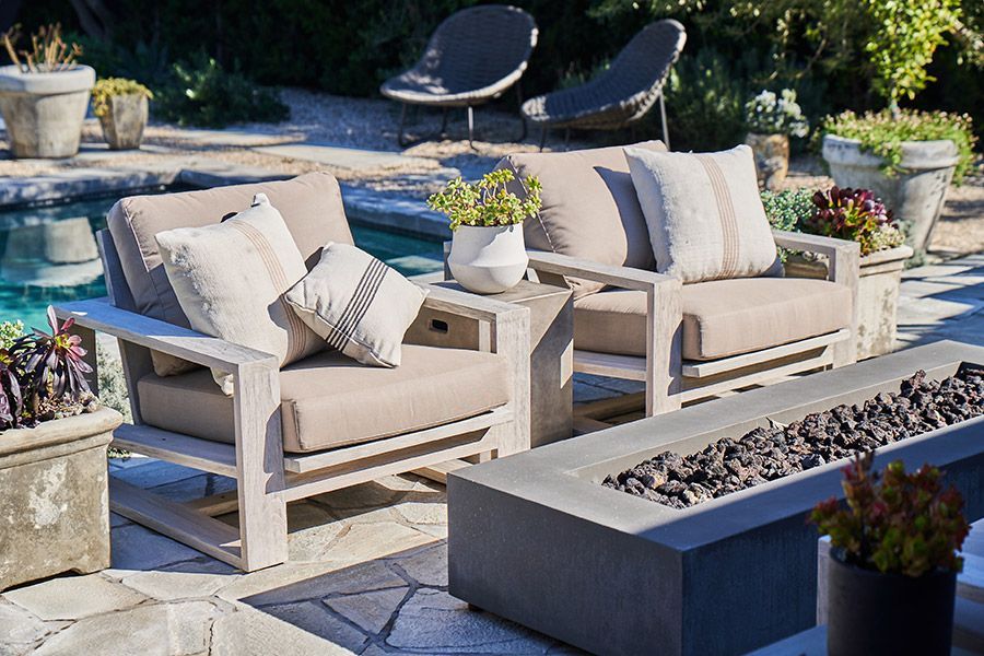 Two light-wood patio chairs with beige cushions and patterned pillows arranged around a long rectangular fire pit.