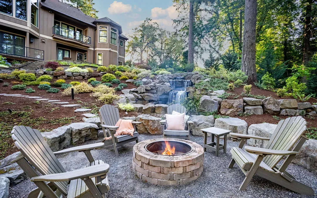 A rustic fire pit area with Adirondack chairs, set in a landscaped backyard with a stone retaining wall and waterfall.