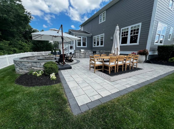 A gray house exterior with a stone patio featuring an outdoor dining set, an umbrella, and a fire pit area.