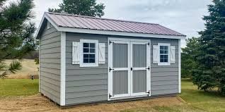 A gray wooden shed with white trim, double doors, and a reddish metal roof, situated on a grassy lawn with trees.