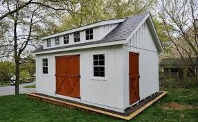 A white shed with brown double doors, a side door, and a row of transom windows, set on a wooden base in a grassy yard.