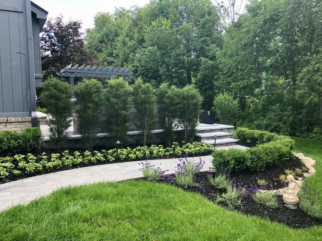 A stone walkway leads toward a patio area with a pergola and manicured hedges in a lush, green backyard.