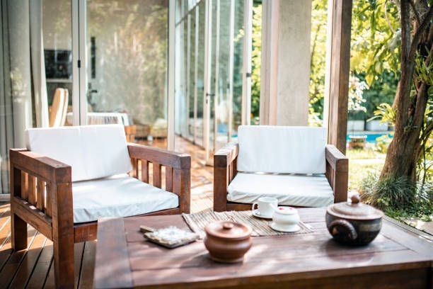 Two wooden chairs with white cushions face a low wooden coffee table with tea service on a deck near a lush garden.