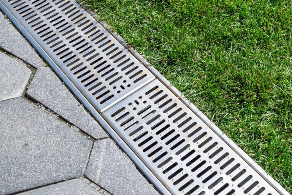 A silver metal trench drain grate installed between a gray stone paver patio and a green lawn.