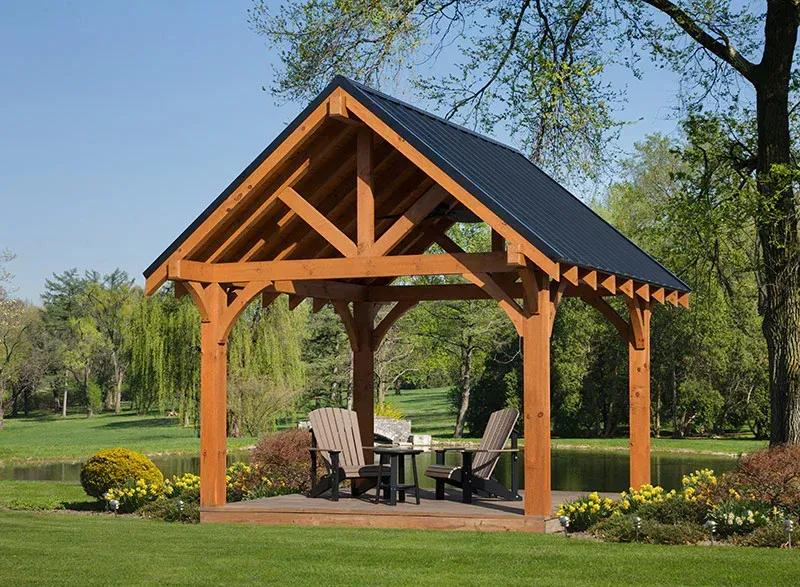 A timber-frame gazebo with a black metal roof sits on a deck by a pond, featuring two chairs and a small table.