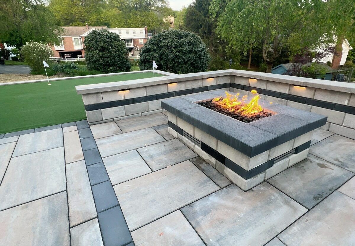 A modern stone fire pit on a paver patio next to a putting green and a low stone seating wall with integrated lighting.