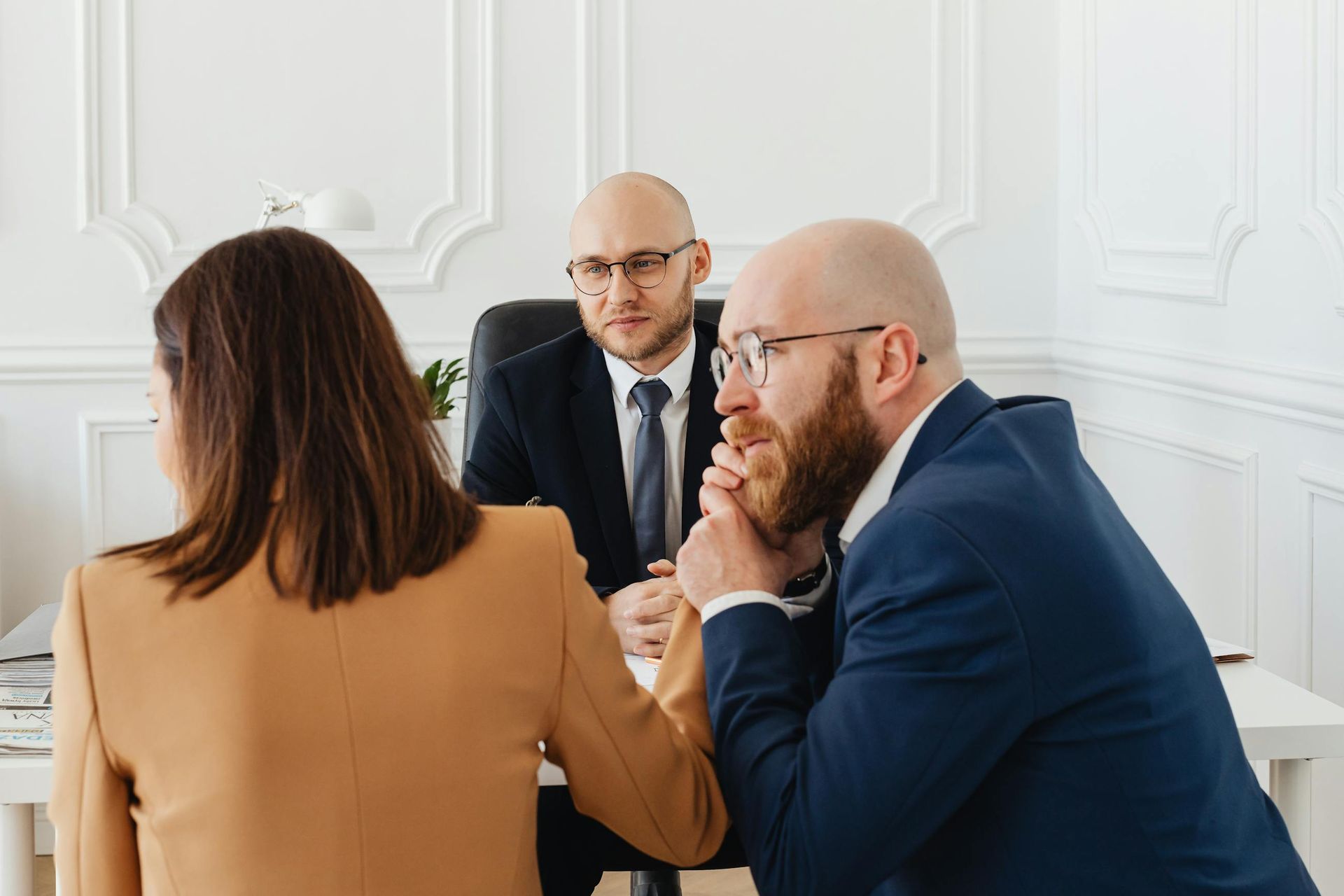 Three people in a business meeting, seated around a table in a bright office, one man speaking thoughtfully