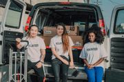 Three people sit in the back of an open van with boxes, wearing matching shirts, smiling at the camera.