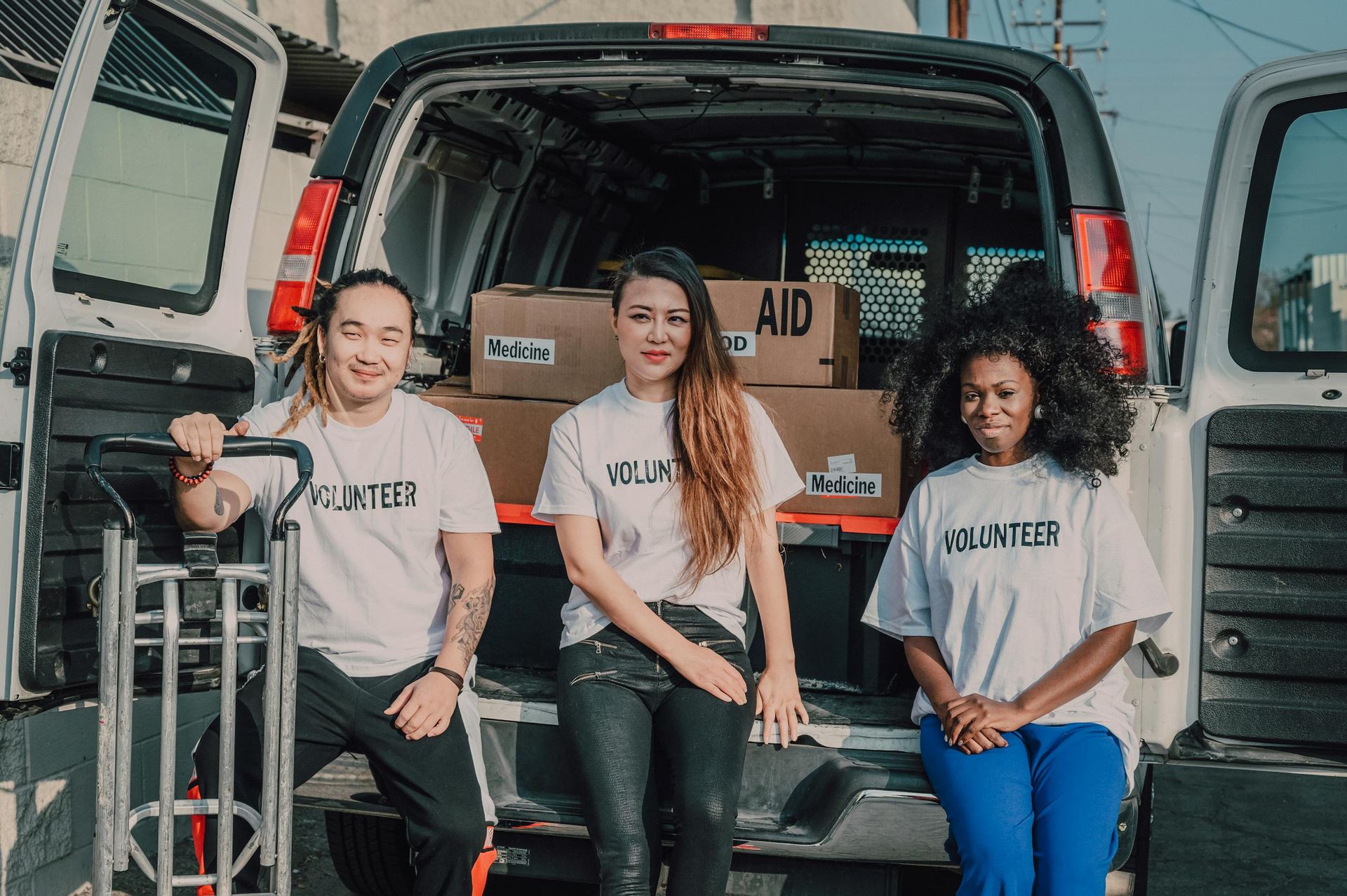 Three people sit in the back of an open van with boxes, wearing matching shirts, smiling at the camera.