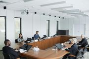 Conference room meeting with several people seated around a large wooden table, laptops open and a projector overhead