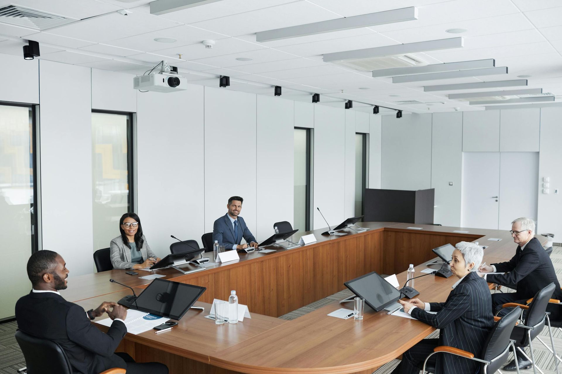 Conference room meeting with several people seated around a large wooden table, laptops open and a projector overhead