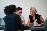 Three people in a meeting discussing papers at a table, with a laptop in the background.