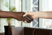 Two hands shaking in a bright indoor setting, one dark-skinned and one light-skinned.