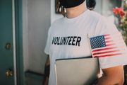 Person in a white “VOLUNTEER” shirt holding a folder and small American flag at a doorway