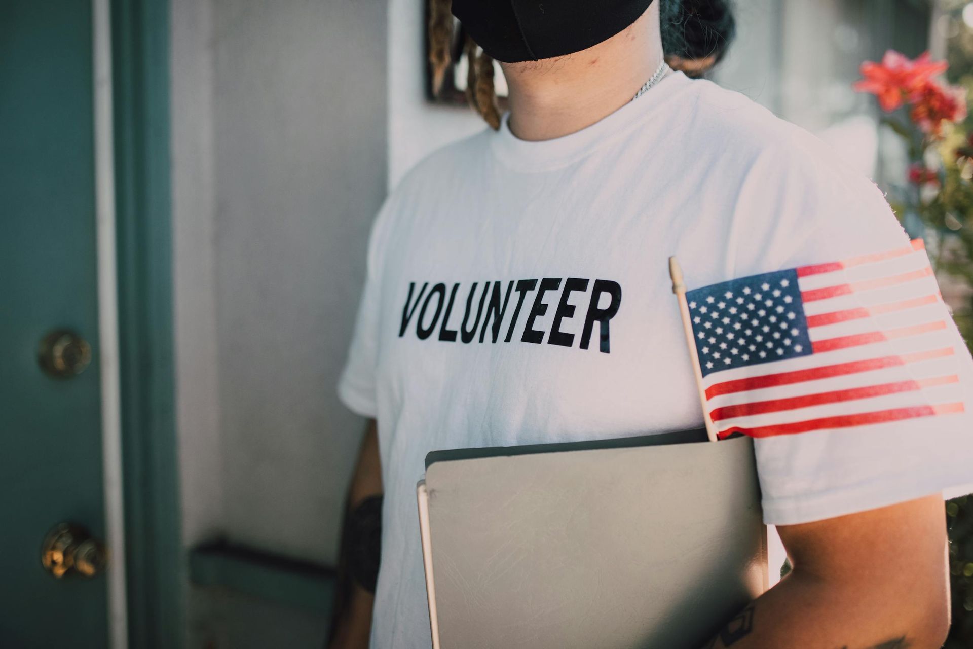 Person in a white “VOLUNTEER” shirt holding a folder and small American flag at a doorway