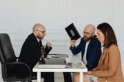 Three people in an office meeting, one holding up a tablet while two others sit across the desk.