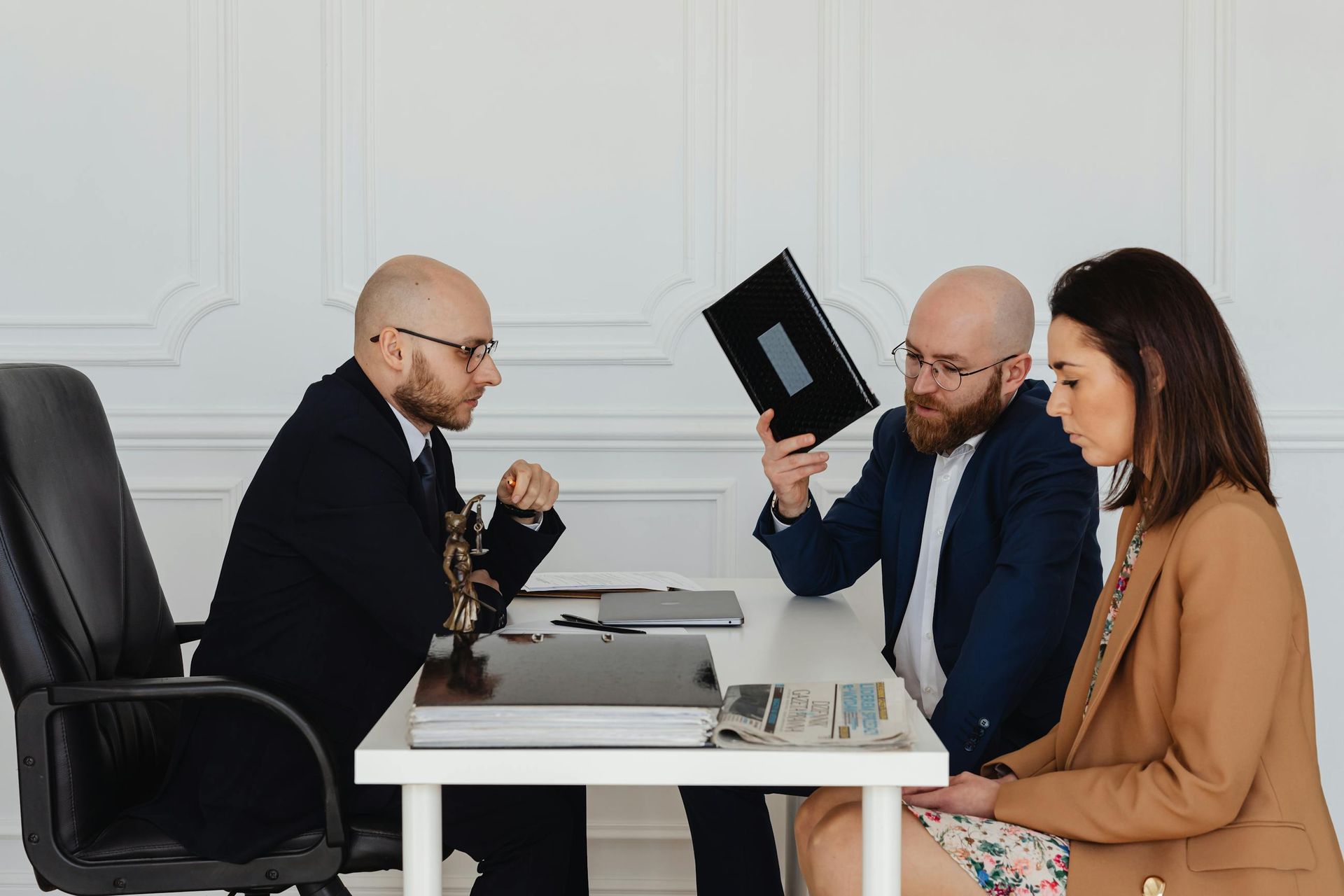 Three people in an office meeting, one holding up a tablet while two others sit across the desk.